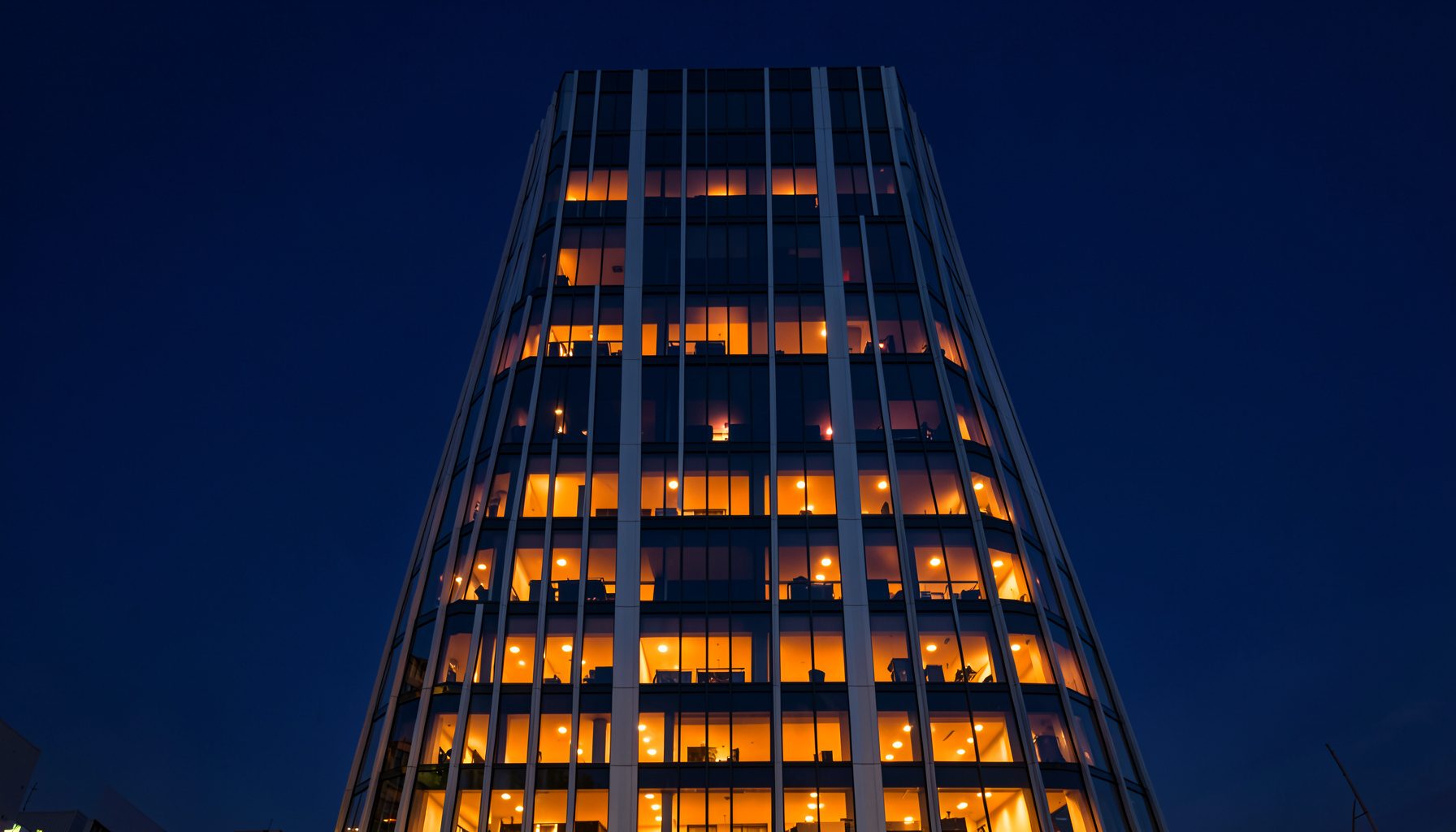 A symmetrical wide-angle view of a modern glass-and-steel office tower facade at dusk, deep navy sky reflected in the windows, warm amber interior lights glowing through, clean vertical lines and strong geometric balance.